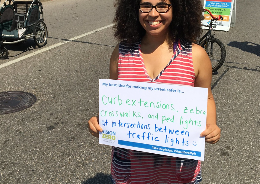 Person holding sign saying "Curb extensions, zebra crosswalks and ped lights at intersections between lights." with a smiley face drawn on sign.