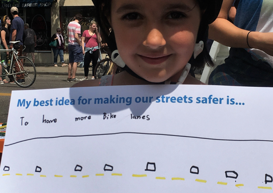 Child wearing helmet holding sign that says "My best idea for making our streets safer is "to have more bike lanes" with drawing of a road.