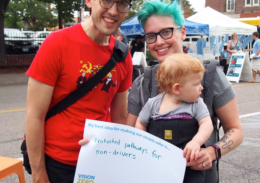 Two adults and a baby. Adult is holding sign that says: "My best idea for making our streets safer. Protected pathways for non-drivers."