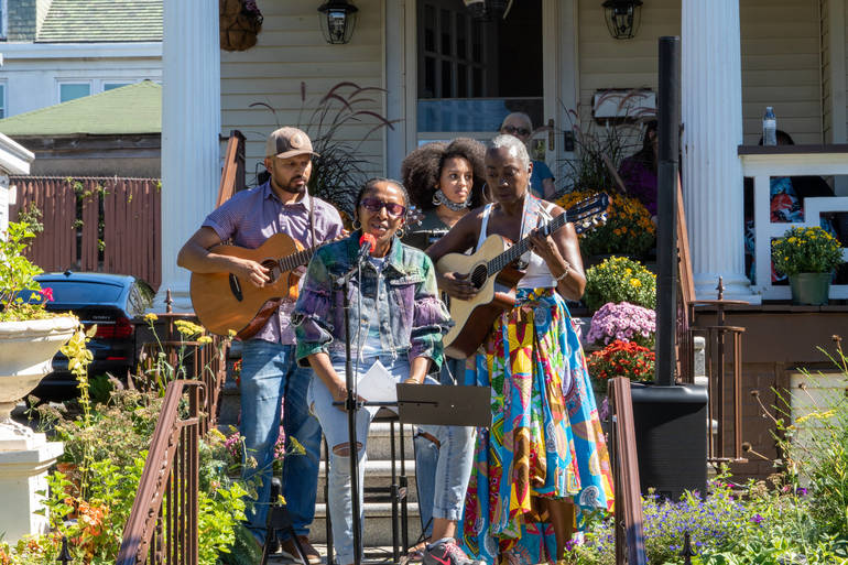 Four musicians perform outdoors on a flower-filled porch, with two acoustic guitars and a singer at a microphone
