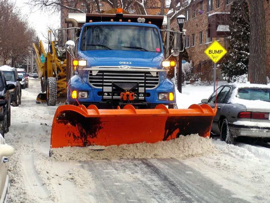 Blue snowplow with orange shovel on front clearing snow from a city street