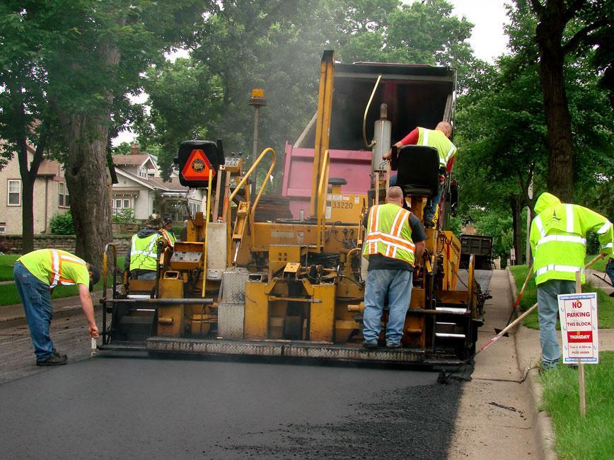 Three people in yellow construction vests operating a large machine to lay fresh asphalt on a street