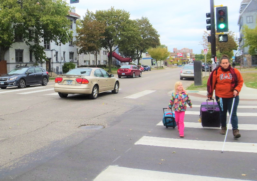 Person with white walking cane and a child, both pulling suitcases. They're crossing at a crosswalk in Minneapolis, MN.